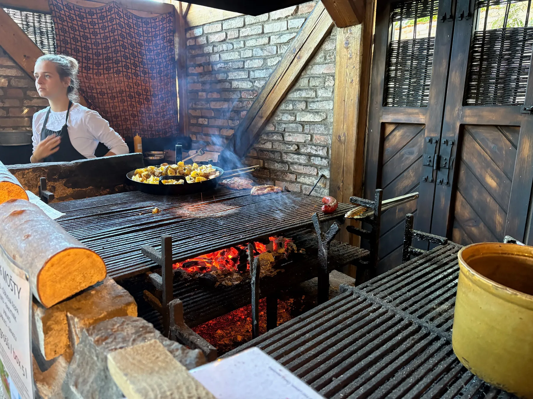 Kuchař u otevřeného grilu se dvěma velkými pánvemi masa a zeleniny, cihlová stěna v pozadí