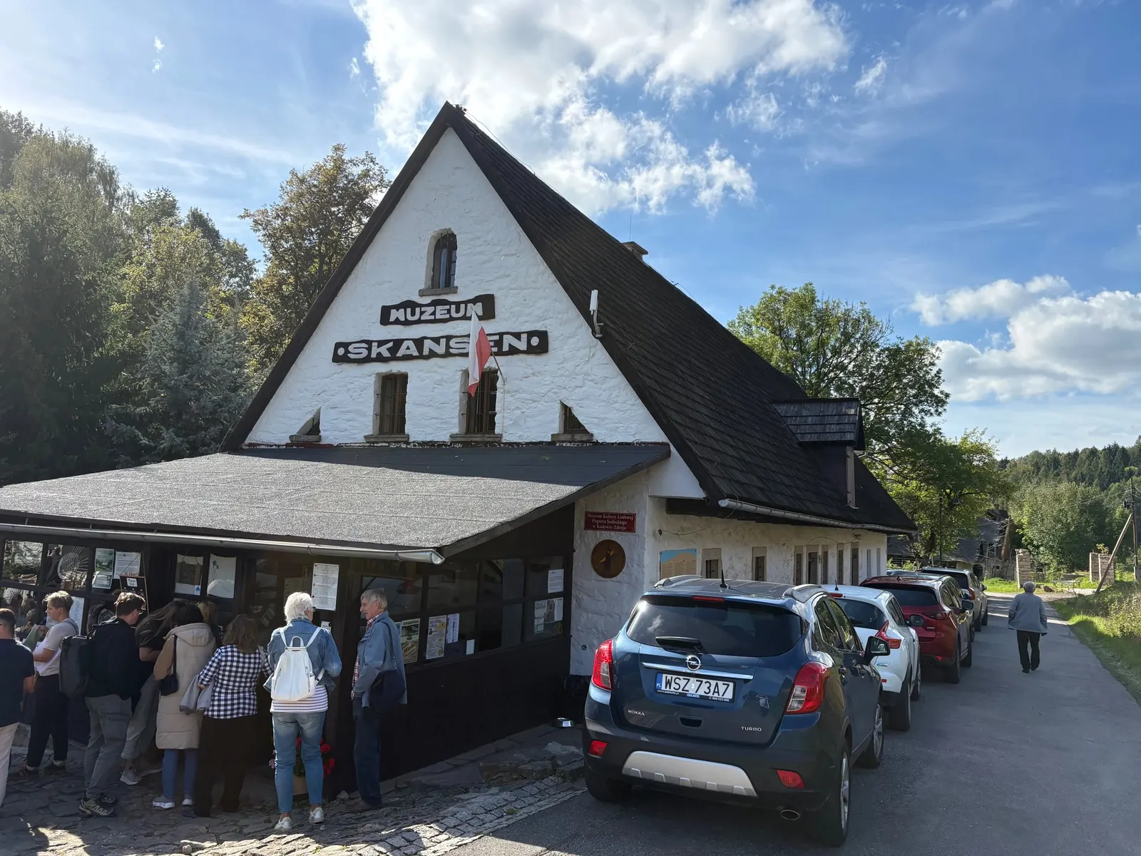 Vstup do skanzenu ve Pstrążné - bílá roubenka s tmavou sedlovou střechou, nápis MUZEUM SKANSEN, u cesty parkující auta