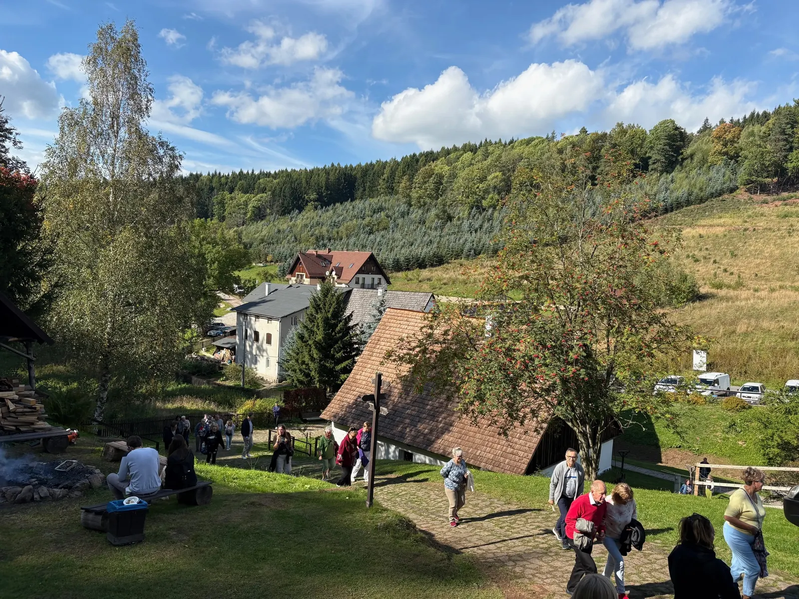 Panorama skanzenu - roubenky, ohniště se stoupajícím kouřem, návštěvníci, kopce v pozadí