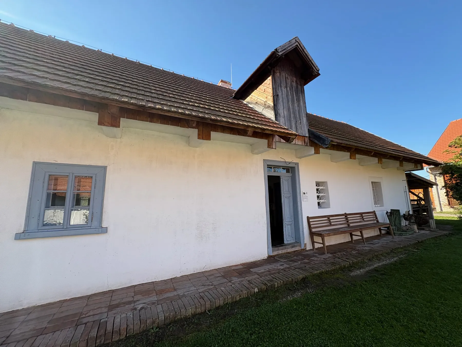 Bílá chalupa s cihlovou střechou, modré okenice a modrý dveřní rám, dřevěný komín