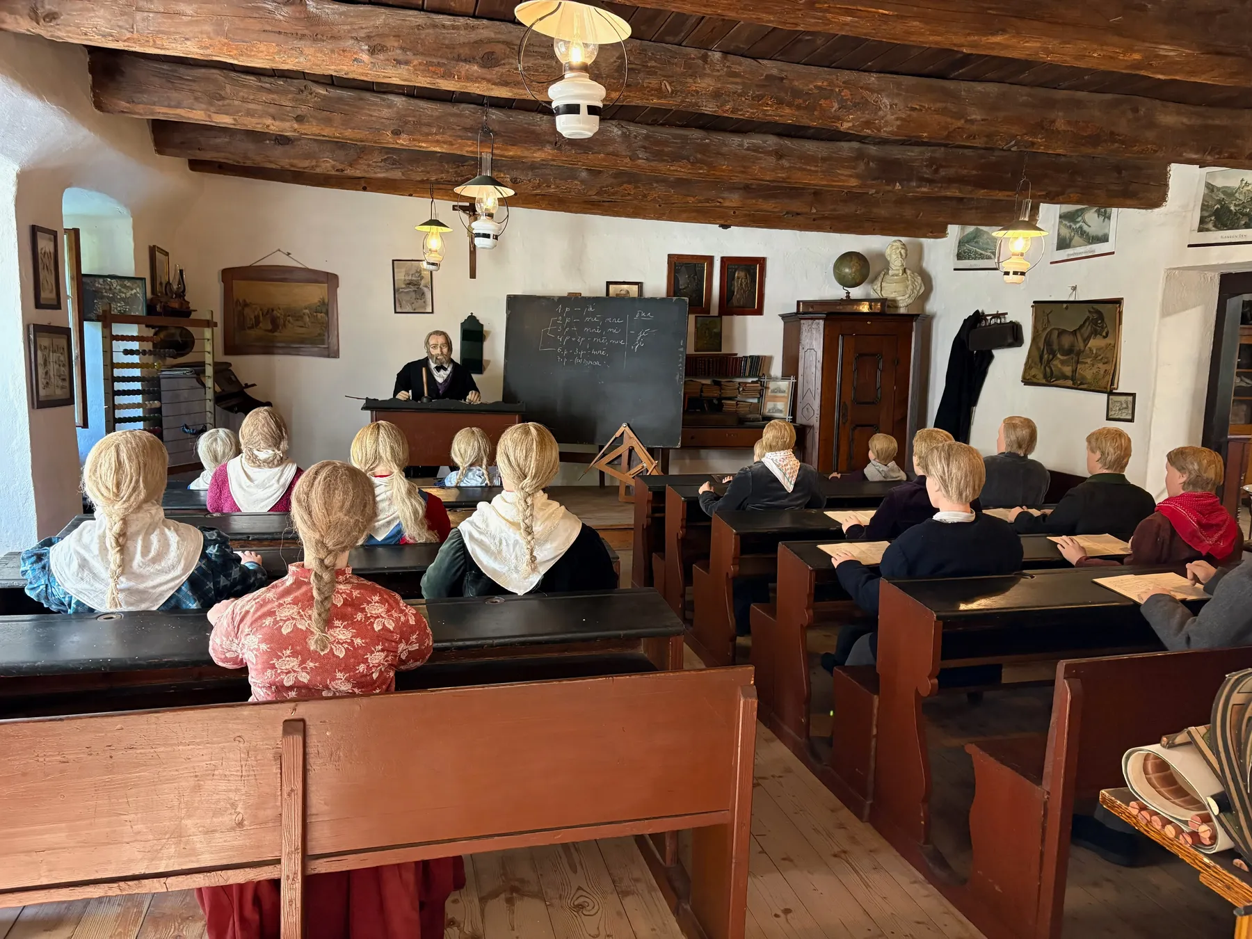 Vnitřek historické školy - žáci figuríny v lavicích, učitel u tabule, dřevěné trámy stropu