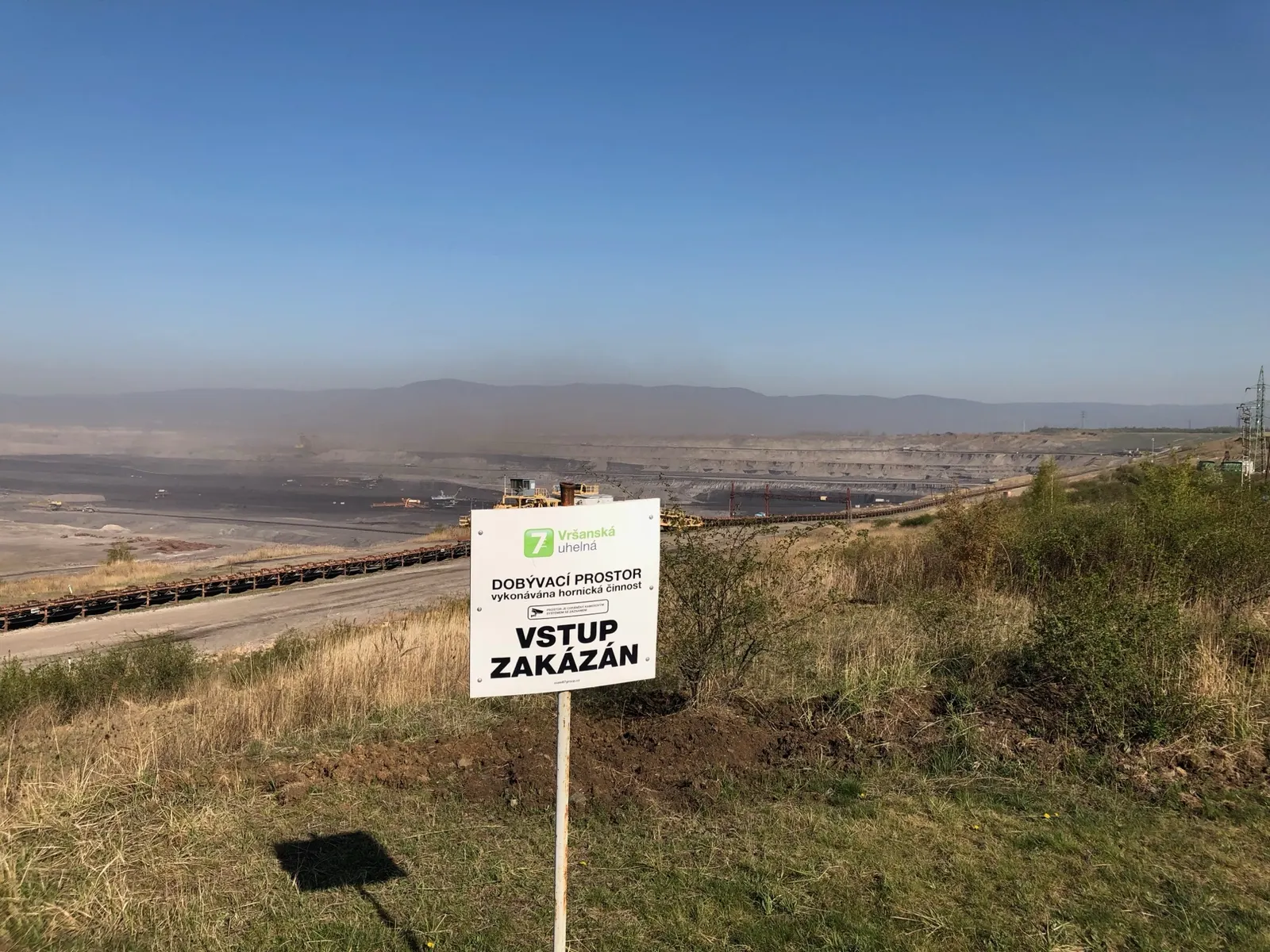 Panoramatický pohled na dobývací prostor Vršan z vyhlídkové terasy, s cedulí Vršanské uhelné a Krušnými horami v pozadí